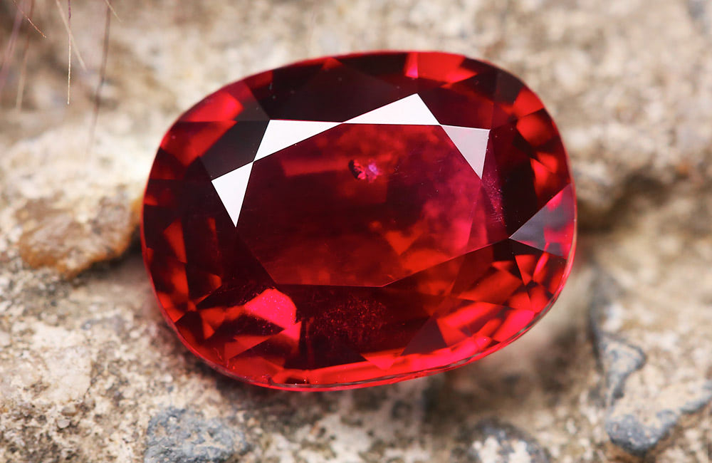 A faceted red gemstone lies on a rough, stone surface, displaying a vibrant, reflective brilliance amidst an earthy background.