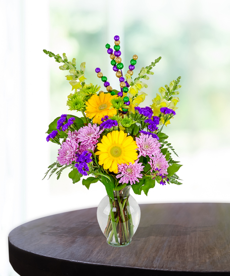 A Mardi Gras arrangement featuring yellow gerbera daisies, lavender mums, green button poms, and more  in a clear glass vase with colorful Mardi Gras accents.