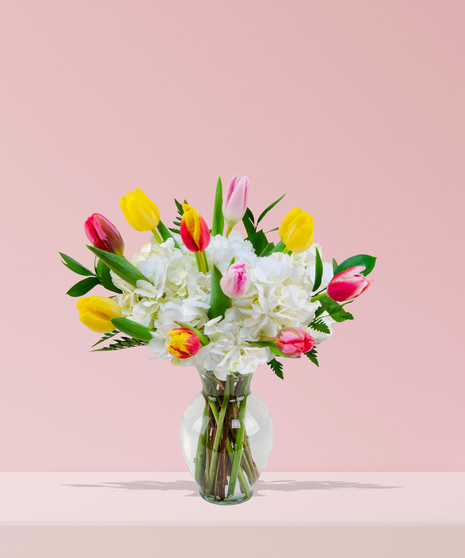 A vibrant bouquet of colorful tulips in yellow, orange, pink, and red, paired with white hydrangeas and greenery in a clear glass vase.