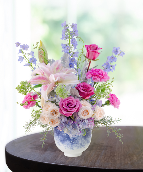 Pastel floral arrangement featuring lilies, roses, carnations, hydrangeas, and delphiniums in a lavender vase.