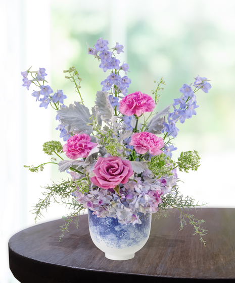 Pastel floral arrangement featuring lilies, roses, carnations, hydrangeas, and delphiniums in a lavender vase.