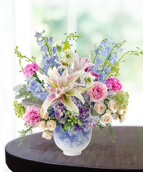 Pastel floral arrangement featuring lilies, roses, carnations, hydrangeas, and delphiniums in a lavender vase.