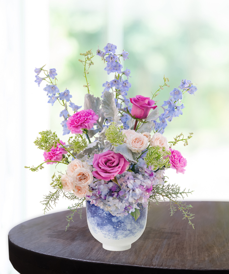 Pastel floral arrangement featuring lilies, roses, carnations, hydrangeas, and delphiniums in a lavender vase.