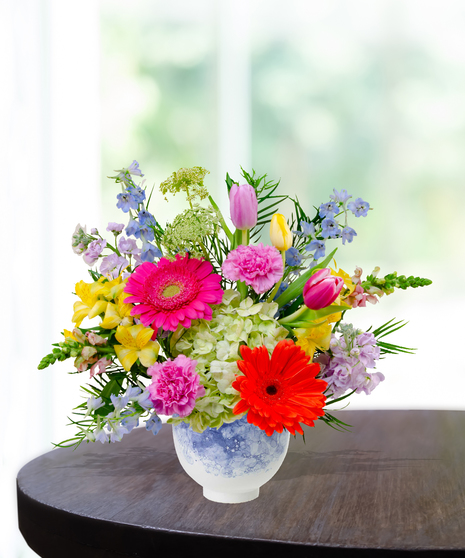 Hummingbird Hues floral arrangement with peach garden roses, green hydrangeas, gerbera daisies, and mixed colorful blooms in a blue-and-white patterned vase on a wooden table.