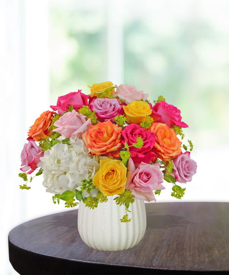 Groovy Garden floral arrangement with pink, yellow, orange, and lavender roses, white hydrangeas, and fresh greenery in a white textured vase.