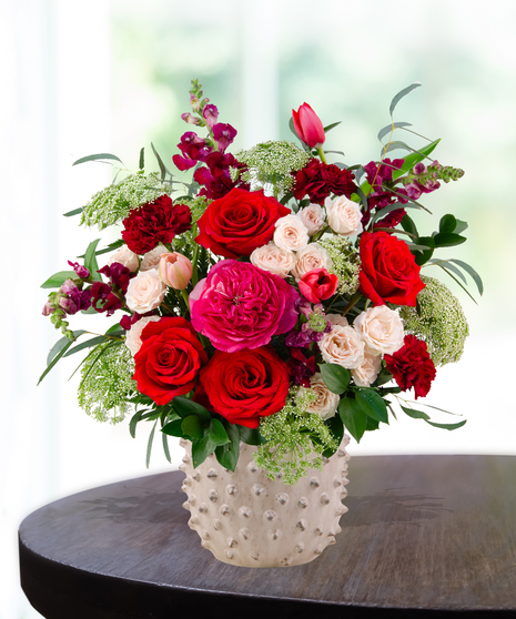 Garnet N' Roses floral arrangement with red roses, fuchsia garden roses, blush spray roses, and burgundy snapdragons in a textured white ceramic vase.