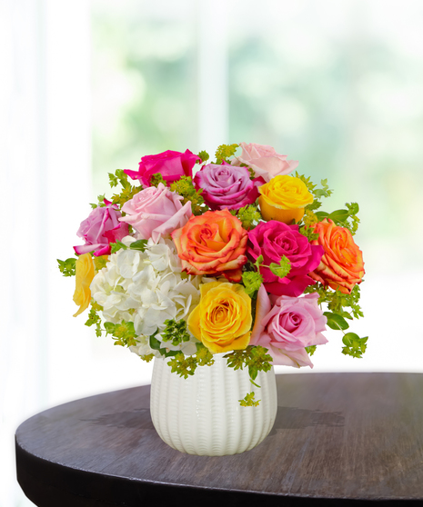 Groovy Baby floral arrangement with pink, yellow, orange, and lavender roses, white hydrangeas, and fresh greenery in a white textured vase.