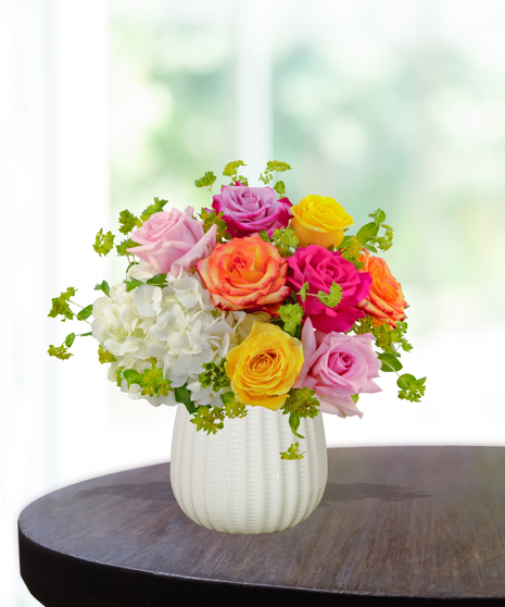 Groovy Garden floral arrangement with pink, yellow, orange, and lavender roses, white hydrangeas, and fresh greenery in a white textured vase.