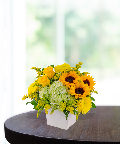 Lemon Drop floral arrangement with sunflowers, yellow roses, green hydrangeas, and solidago in a modern white vase on a wooden table.