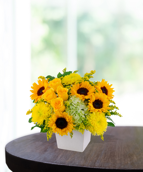 Lemon Drop floral arrangement with sunflowers, yellow roses, green hydrangeas, and solidago in a modern white vase on a wooden table.