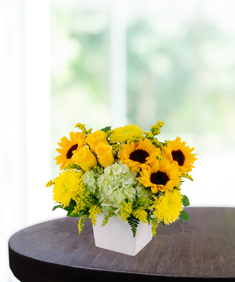 Lemon Drop floral arrangement with sunflowers, yellow roses, green hydrangeas, and solidago in a modern white vase on a wooden table.