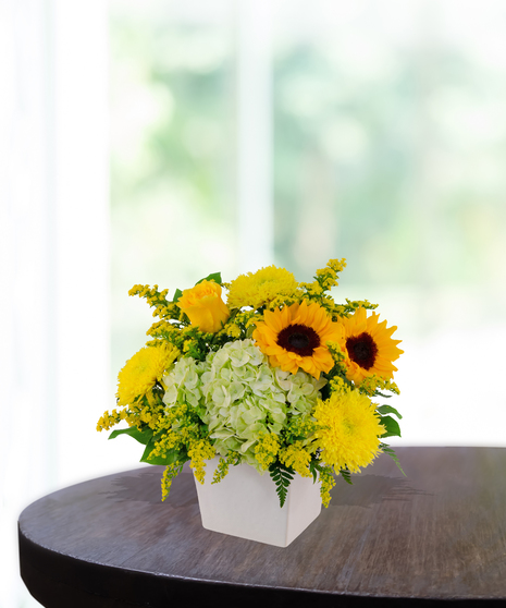 Lemon Drop floral arrangement with sunflowers, yellow roses, green hydrangeas, and solidago in a modern white vase on a wooden table.