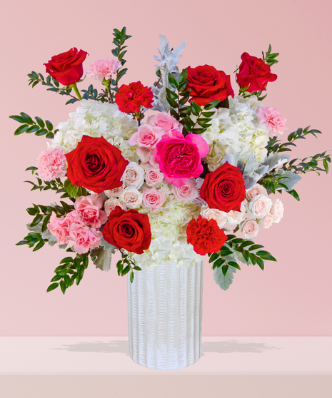 P.S. I Love You Valentine’s Day floral arrangement with red roses, fuchsia garden roses, pink spray roses, and carnations in a white textured vase.