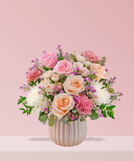 Love Story Valentine’s Day floral arrangement with pink and peach roses, fuchsia garden blooms, carnations, and lavender asters in a white textured vase.