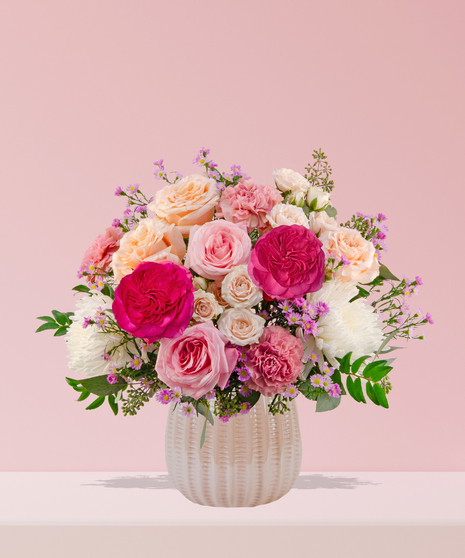 Love Story Valentine’s Day floral arrangement with pink and peach roses, fuchsia garden blooms, carnations, and lavender asters in a white textured vase.