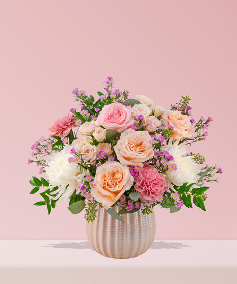 Love Story Valentine’s Day floral arrangement with pink and peach roses, fuchsia garden blooms, carnations, and lavender asters in a white textured vase.