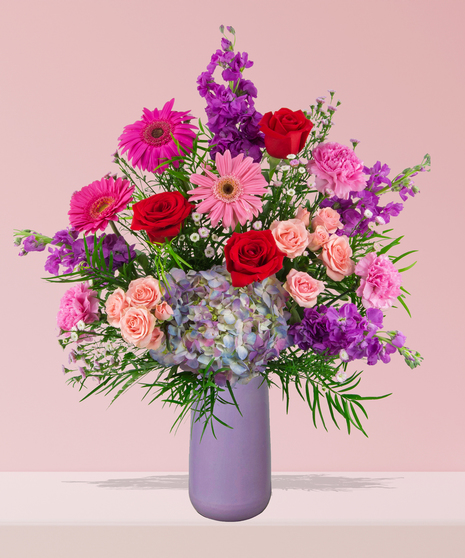 A vibrant flower arrangement featuring red roses, pink gerbera daisies, and more in a lavender vase, exclusively from Billy Heroman’s Florist in Baton Rouge, LA.