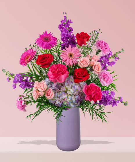 A vibrant flower arrangement featuring red roses, pink gerbera daisies, and more in a lavender vase, exclusively from Billy Heroman’s Florist in Baton Rouge, LA.