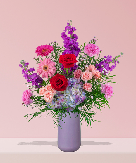 A vibrant flower arrangement featuring red roses, pink gerbera daisies, and more in a lavender vase, exclusively from Billy Heroman’s Florist in Baton Rouge, LA.