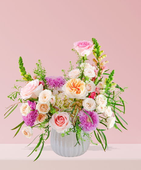 Smitten floral arrangement with peach and pink roses, lavender mums, and snapdragons in a white textured vase.