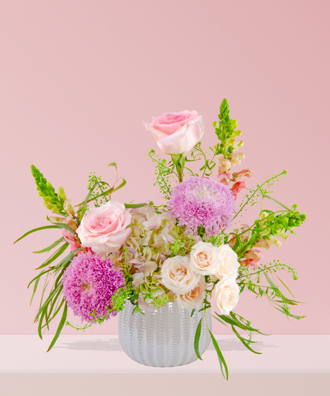 Smitten floral arrangement with peach and pink roses, lavender mums, and snapdragons in a white textured vase.