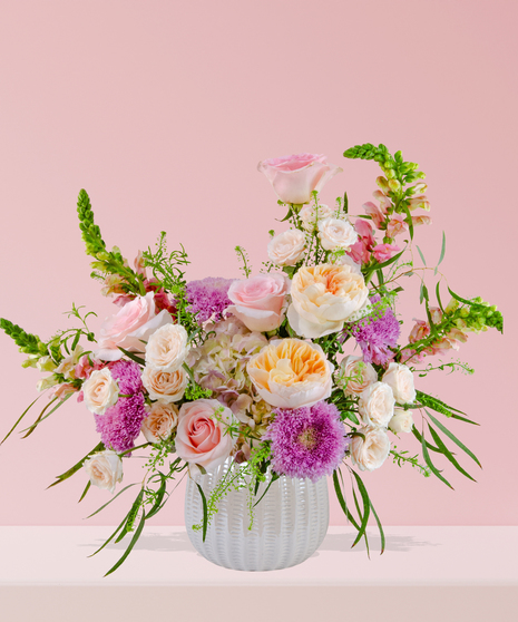 Smitten floral arrangement with peach and pink roses, lavender mums, and snapdragons in a white textured vase.