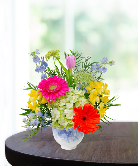 Hummingbird Hues floral arrangement with peach garden roses, green hydrangeas, gerbera daisies, and mixed colorful blooms in a blue-and-white patterned vase on a wooden table.