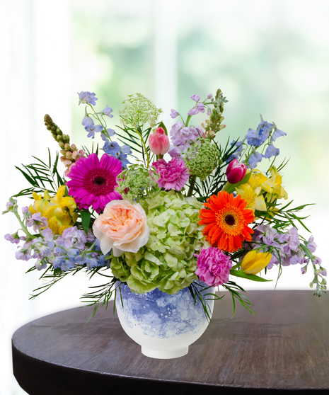 Hummingbird Hues floral arrangement with peach garden roses, green hydrangeas, gerbera daisies, and mixed colorful blooms in a blue-and-white patterned vase on a wooden table.