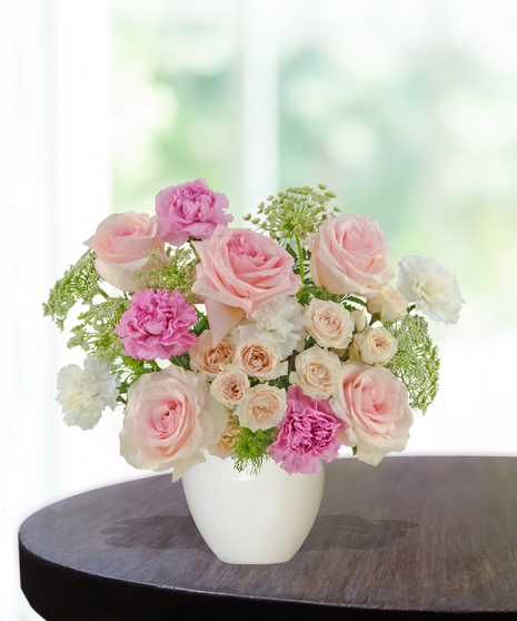 A pastel floral arrangement featuring pink roses, spray roses, and carnations, accented with greenery and Queen Anne’s lace in a white vase by Billy Heroman’s.