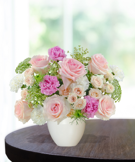 A pastel floral arrangement featuring pink roses, spray roses, and carnations, accented with greenery and Queen Anne’s lace in a white vase by Billy Heroman’s.