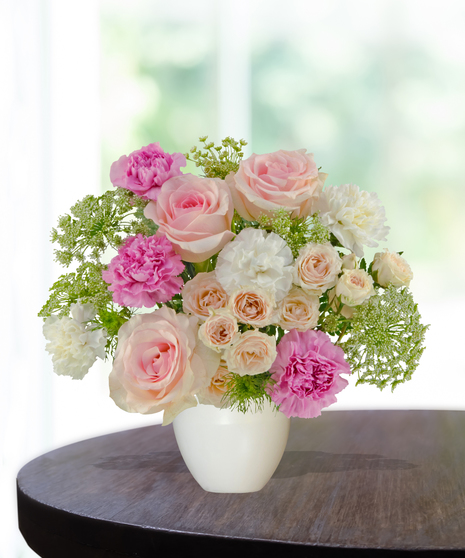 A pastel floral arrangement featuring pink roses, spray roses, and carnations, accented with greenery and Queen Anne’s lace in a white vase by Billy Heroman’s.