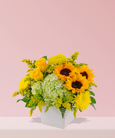 Vibrant floral arrangement with yellow sunflowers, roses, hydrangeas, chrysanthemums, and greenery in a white ceramic vase.