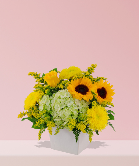 Vibrant floral arrangement with yellow sunflowers, roses, hydrangeas, chrysanthemums, and greenery in a white ceramic vase.