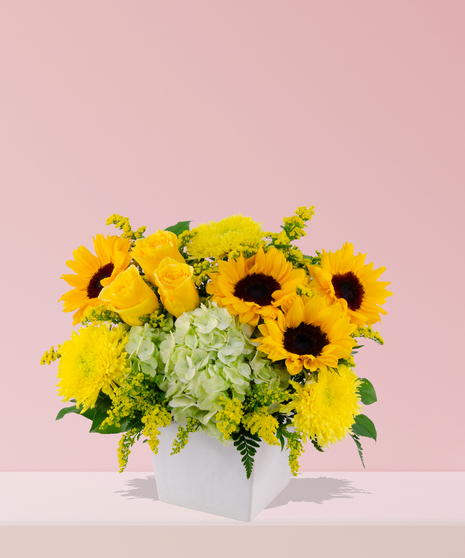 Vibrant floral arrangement with yellow sunflowers, roses, hydrangeas, chrysanthemums, and greenery in a white ceramic vase.