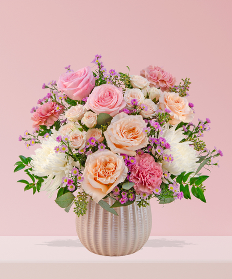 Love Story Valentine’s Day floral arrangement with pink and peach roses, fuchsia garden blooms, carnations, and lavender asters in a white textured vase.