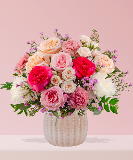 Love Story Valentine’s Day floral arrangement with pink and peach roses, fuchsia garden blooms, carnations, and lavender asters in a white textured vase.