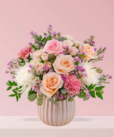 Love Story Valentine’s Day floral arrangement with pink and peach roses, fuchsia garden blooms, carnations, and lavender asters in a white textured vase.