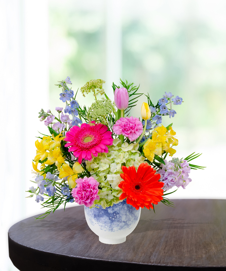 Hummingbird Hues floral arrangement with peach garden roses, green hydrangeas, gerbera daisies, and mixed colorful blooms in a blue-and-white patterned vase on a wooden table.