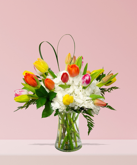 A vibrant bouquet of colorful tulips in yellow, orange, pink, and red, paired with white hydrangeas and greenery in a clear glass vase.