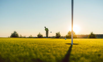 A golfer putts on a green under a clear sky at sunset, with surrounding trees casting long shadows.