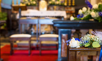 A wooden bench adorned with white and blue flowers sits in the foreground of a church interior, featuring a red carpet and an altar decorated with flowers in the background.