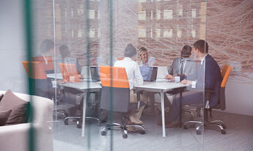 A group of professionals sits around a conference table, discussing or collaborating, inside a modern glass-walled office with a view of a cityscape through the windows.