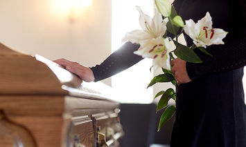 A person holding white lilies rests a hand on a wooden casket in a softly lit room, suggesting a funeral or memorial service.